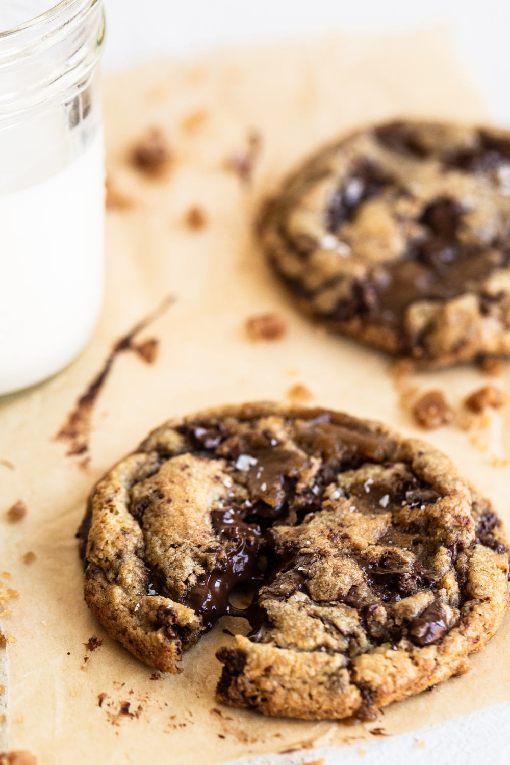two Browned Butter Toffee Chocolate Chip Cookies on parchment paper with a glass of milk