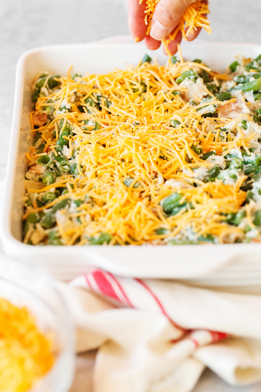 a hand sprinkling cheese across the Green Bean Casserole in its white ceramic casserole dish, before baking.