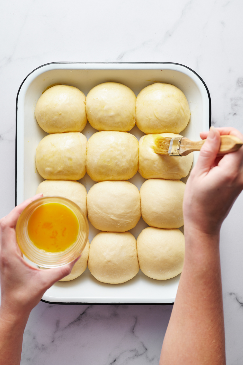 a pan of dinner rolls being brushed with egg wash before baking.